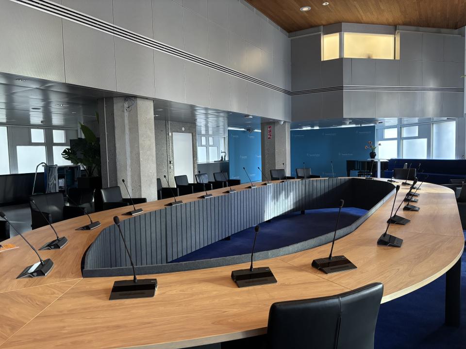 Een ovale tafel met een open ruimte in het midden. Het blad is in houtkleur en er staan een aantal microfoons opgesteld.