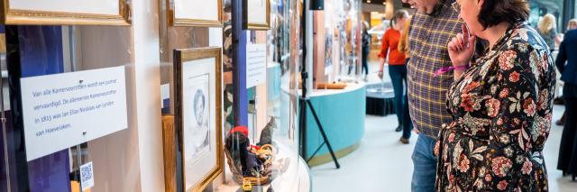 Een man en een vrouw rechts in beeld staan voor een staande vitrine, waarin documenten en portretten zichtbaar zijn. 