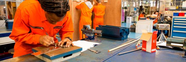 Jongeren in oranje overalls. Vooraan buigt een van hen zich over een werktafel, daarachter staan twee studenten met de rug naar de camera bij een houten scheidingswand.