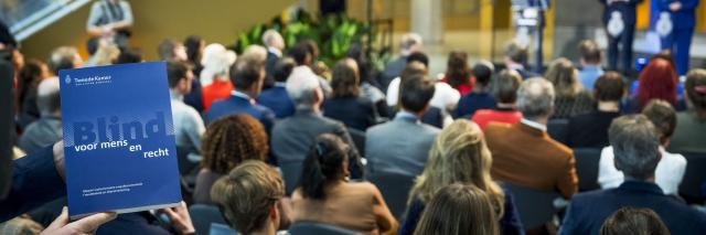 Op de voorgrond houdt een hand het rapport Blind voor mens en recht vast. Op de achtergrond de Statenpassage met toeschouwers die zitten op stoelen en een podium waarop drie commissieleden staan.