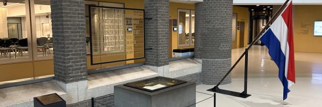 Een stenen blok met daarop een glasplaat. Onder het glas is een opengeslagen boek zichtbaar, dat is de Erelijst van Gevallenen. Rechts daarvan de Nederlandse vlag in een houder en op de achtergrond glazen wanden. Daarachter liggen de zalen voor commissievergaderingen. 