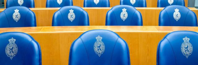 Overzicht van vier rijen blauwe zetels achter de Kamerbankjes in de plenaire zaal van de Tweede Kamer. 