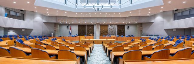 Plenaire zaal leeg, vanachter de Kamerzetels gezien. In het plafond is een groot lichtarmatuur te zien. 