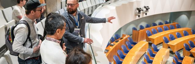Een man wijst een groepje bezoekers vanaf de publieke tribune onderdelen van de plenaire zaal aan. 