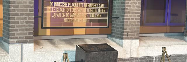 Stenen plaquette op een stenen zuil, met ervoor standaards voor kransen. Erachter een uitleg in tekst over de plaquette. 
