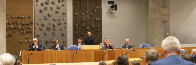 In de plenaire zaal van de Tweede Kamer is in de verte president Zelensky zichtbaar, hij staat achter het spreekgestoelte waar normaal gesproken de minister staat. De Kamerleden zijn op de rug zichtbaar. 
