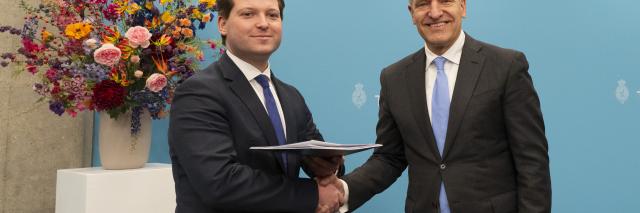 Kamervoorzitter Thom van Campen schudt de hand van informateur Sybrand Buma naast hem en houdt met de andere hand het verslag vast dat hij zojuist heeft gekregen. Beide mannen staan voor een lichtblauwe wand met het Kamerlogo, naast een bloemstuk op een pilaar. 