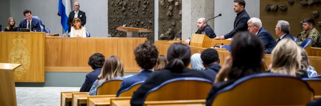 President Zelensky staat achter het katheder en spreekt het parlement toe in de plenaire zaal. Kamerleden zijn zichtbaar op de rug, Kamervoorzitters Thom van Campen en Mei Li Vos zitten op hun plek links op de foto. 