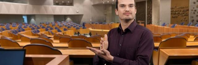 Gebarentolk met daarachter de plenaire zaal van de Tweede Kamer.
