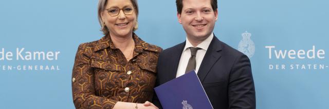 Twee personen staan naast elkaar voor een lichtblauwe wand met logo's van de Tweede Kamer. Links informateur Letschert, rechts Kamervoorzitter Thom van Campen. Zij schudden elkaar de hand en Van Campen houdt het eindverslag van Letschert voor zich. 