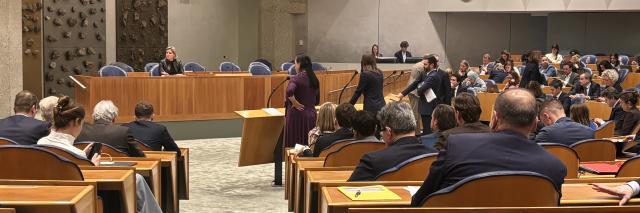 Plenaire vergaderzaal van de Tweede Kamer, de Kamerleden in de zetels zijn op de rug zichtbaar. Op de plek waar normaal gesproken de ministers zitten, vak K, zit nu informateur Rianne Letschert. 