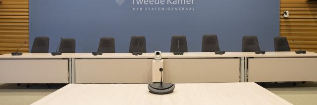 Zaal met op de voorgrond een stoel en tafel met microfoon. Op de achtergrond een tafel met daarachter acht stoelen en een scherm met het logo van de Tweede Kamer.