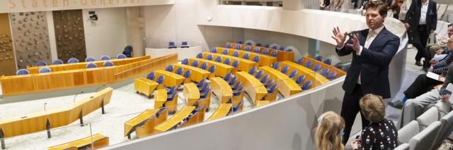Kamervoorzitter Thom van Campen staat op de publieke tribune van de plenaire zaal. Hij vertelt een groep kinderen over het werk van de Tweede Kamer.