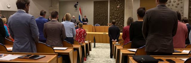 Kamerleden staan in de plenaire zaal bij een herdenking. Zij zijn op de rug gefotografeerd. 