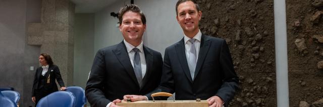 Kamervoorzitter Thom van Campen en minister van Financiën Eelco Heinen poseren samen staand in de plenaire zaal achter de ministerstafel. Ze houden allebei het koffertje met rijksbegroting en Miljoenennota vast. 