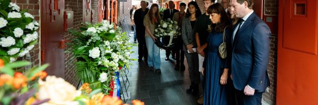 In de gang met gevangeniscellen staan de Voorzitters van Tweede en Eerste Kamer en andere aanwezigen in een lange rij. Zij kijken naar de deuren en de kransen een bloemstukken die neergezet zijn. 