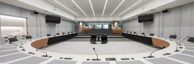 Beeld van lege zaal vanaf de halfronde tafel waaraan tijdens vergaderingen de Kamerleden en bewindspersonen zitten. In de verte is de publieke tribune zichtbaar en daarachter door het raam de Statenpassage. 