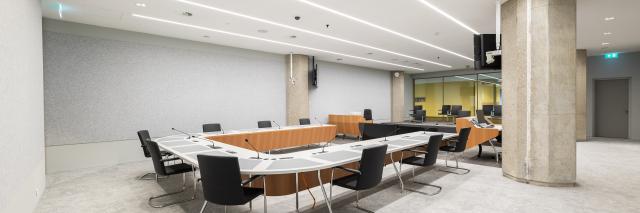 Overzicht van helverlichte witte zaal met in het midden een u-vormige opstelling van tafels met daarachter zwarte stoelen. 