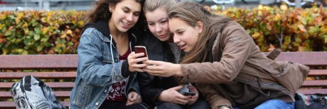 Drie jongeren zitten op een bankje en kijken samen op een telefoon die ze voor zich houden.met social media