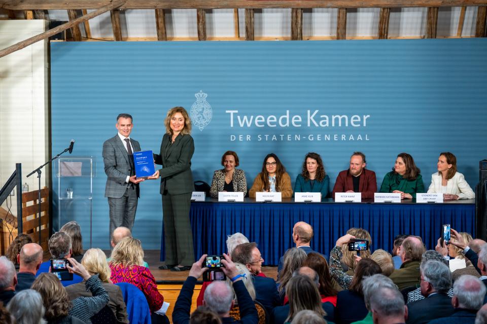 Tom van der Lee overhandigt op een podium het rapport aan Kamervoorzitter Vera Bergkamp.