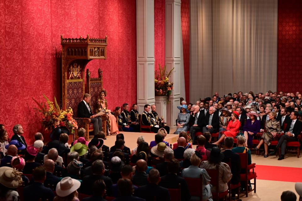 Koning Willem-Alexander leest de troonrede voor, koningin Máxima zit in de zetel ernaast. Zij zitten links op de foto tegen een dieprode wand. Op de voorgrond de leden van Eerste en Tweede Kamer en gasten.