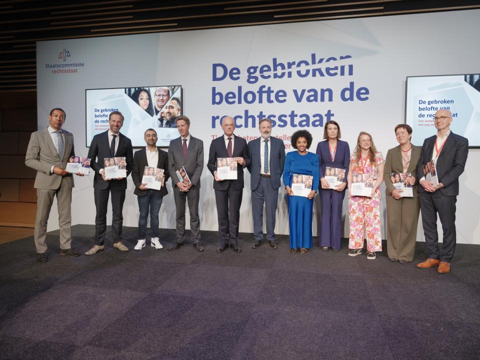 Elf personen staan voor een scherm met de tekst 'Staatscommissie rechtsstaat' en 'De gebroken belofte van de rechtsstaat'. De personen houden in hun hand een document, het adviesrapport van de Staatscommissie rechtsstaat.