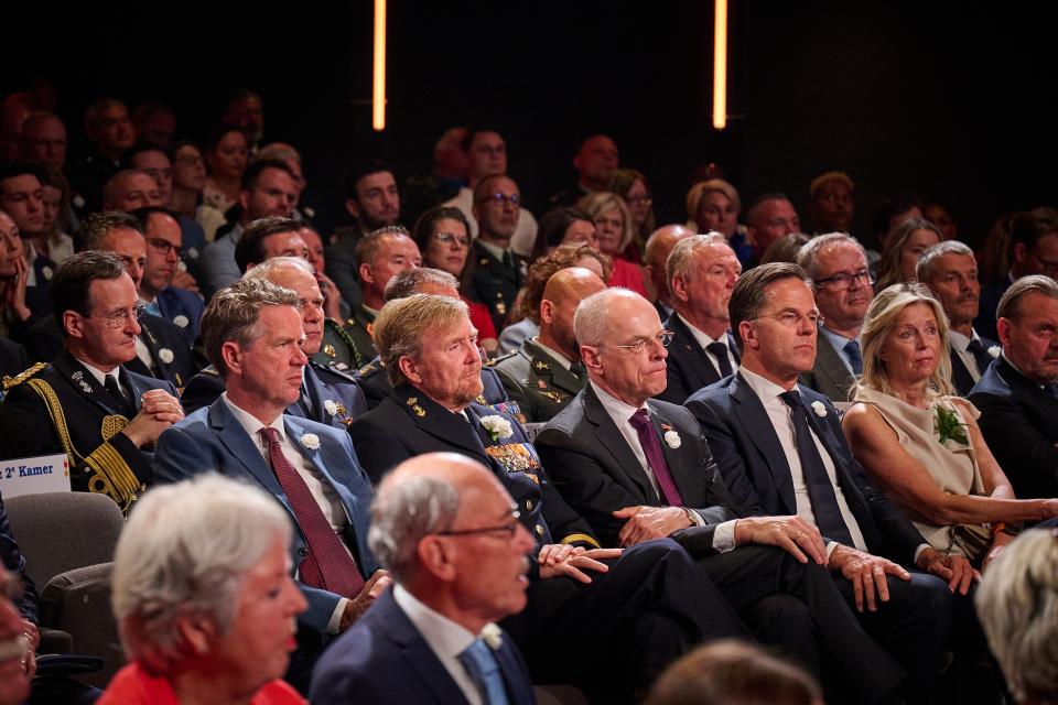 Personen zitten op stoelen in de Koninklijke Schouwburg. Op de voorste rij van links naar rechts Tweede Kamervoorzitter Martin Bosma, Koning Willem-Alexander, Eerste Kamervoorzitter Jan Anthonie Bruijn, demissionair minister-president Mark Rutte en demissionair minister van Defensie Kajsa Ollongren.