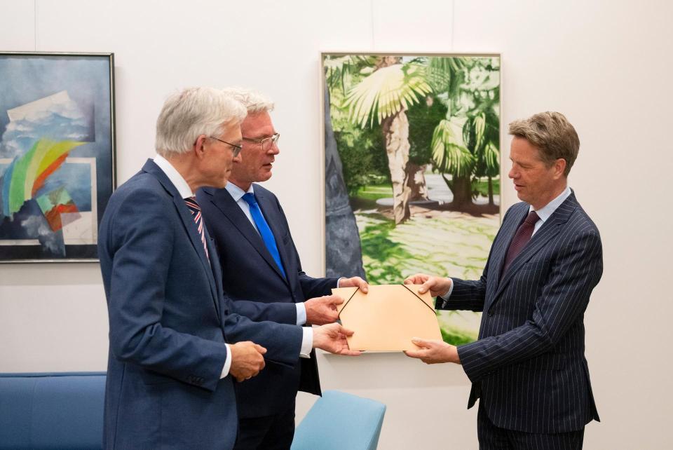 Twee mannen (informateurs Richard van Zwol en Elbert Dijkgraaf) links overhandigen Kamervoorzitter Martin Bosma een document. Ze houden het stuk met zijn drieën vast. Achter hen een wand met verschillende kunstwerken.