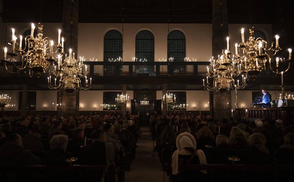 Interieur Portugese Synagoge. Links en rechts grote kroonluchters, daartussen in halfduister publiek op de rug gezien. Rechts in de verte is een spreker uitgelicht.