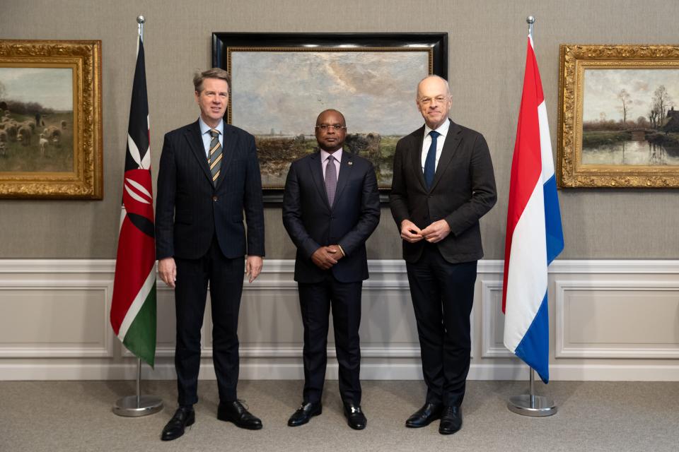 Tussen de Keniase vlag links en de Nederlandse vlag rechts poseren de twee Nederlandse voorzitters met tussen hen in de Keniase collega. Zij staan voor een muur met kunstwerken.