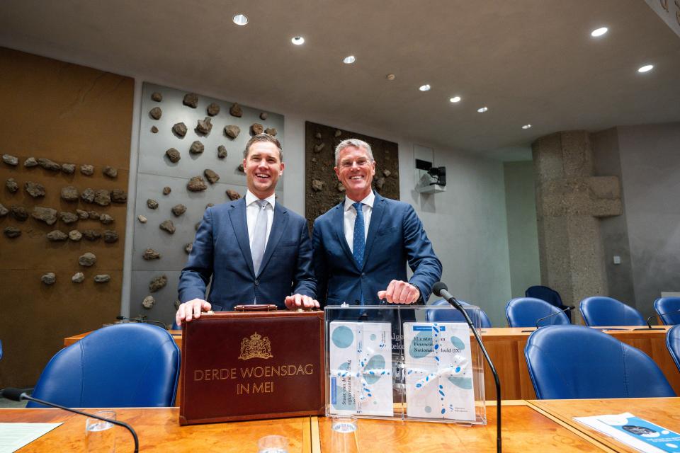 Minister van Financiën Eelco Heinen en president van de Algemene Rekenkamer Pieter Duisenberg staan in vak K van de plenaire zaal. Voor hen staan de twee koffertjes die ze aanbieden. Op de koffer van de minister staat 'Derde woensdag in mei'. De koffer van de Rekenkamer is doorzichtig.