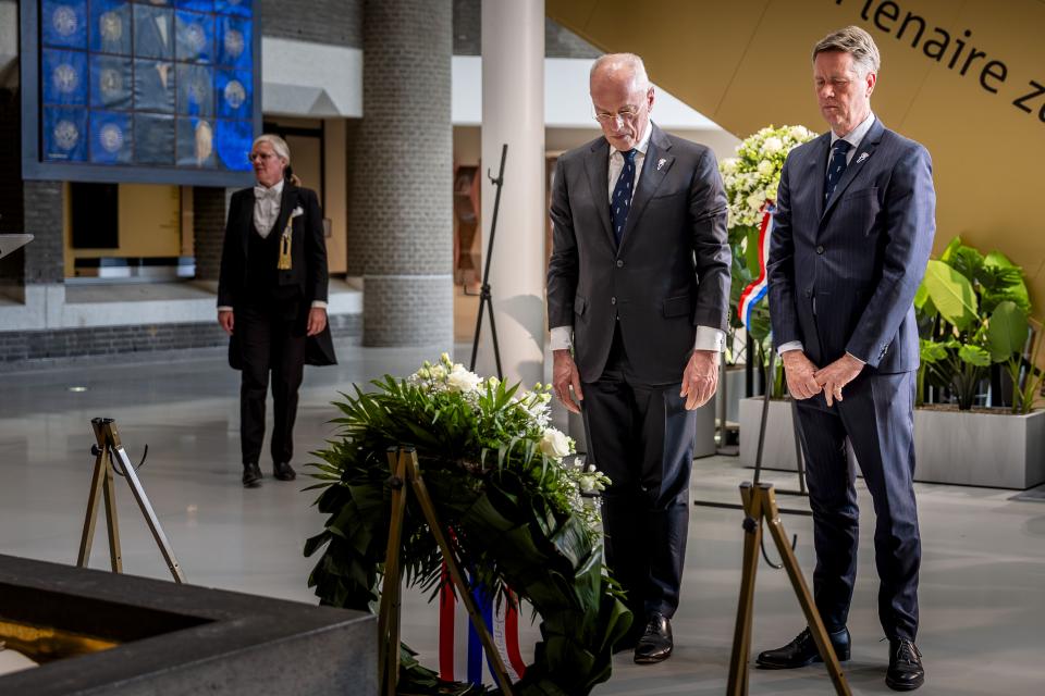De twee Voorzitters staan voor een krans. Je ziet de krans van de achterkant. Ze staan in een lichte ruimte en je ziet een gedeelte van de Erelijst gemaakt van grijze steen. 