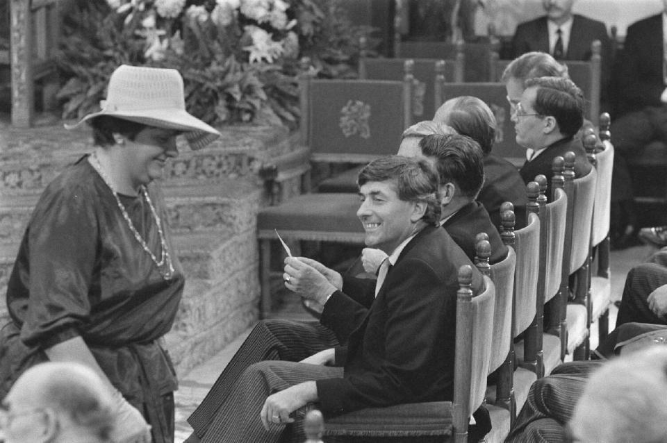 Zwartwit foto uit 1977, een rij stoelen met daarop bewindslieden. VVD-Kamerlid Erica Terpstra met hoed loopt langs.