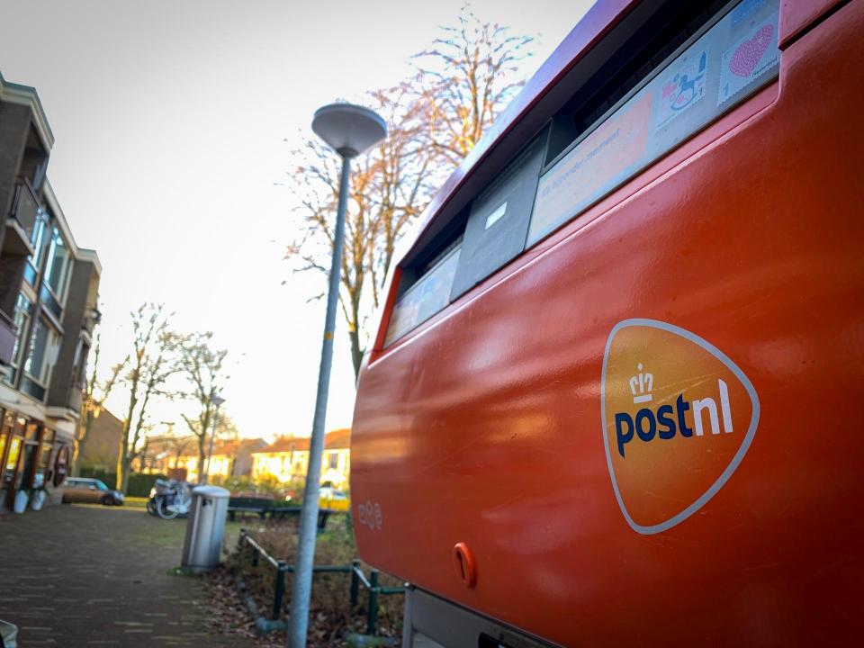 Een rode brievenbus van opzij gezien. De sticker PostNL is zichtbaar. Ernaast een lantaarnpaal en een blok huizen.