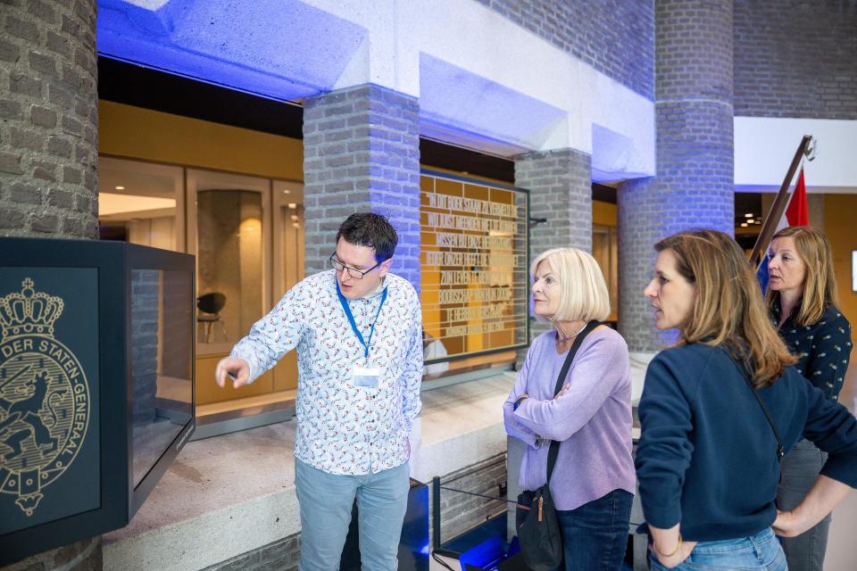 Een medewerker van de Tweede Kamer wijst iets aan op een beeldscherm in het gebouw van de Tweede Kamer, drie bezoekers luisteren toe.