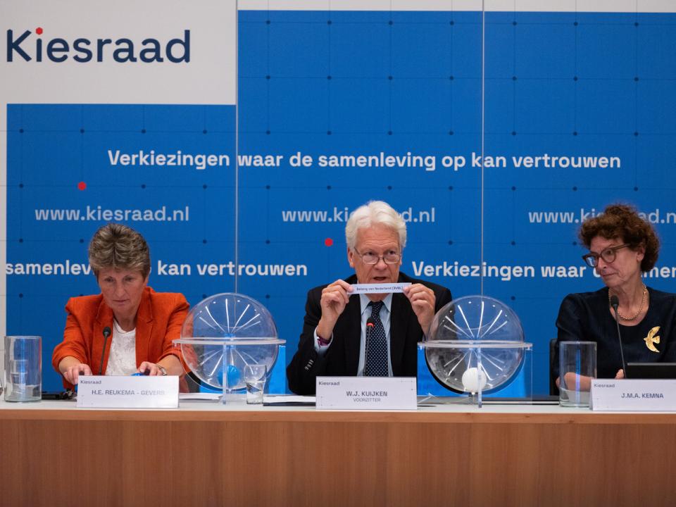 Drie mensen naast elkaar achter een tafel, op de achterwand teksten van de Kiesraad.
