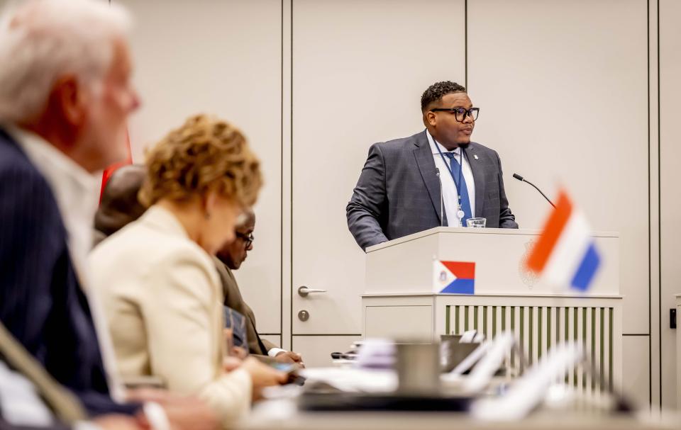 Een man achter het spreekgestoelte in de plenaire zaal van de Eerste Kamer met daarvoor de Nederlandse vlag. Links in beeld toehoorders. 