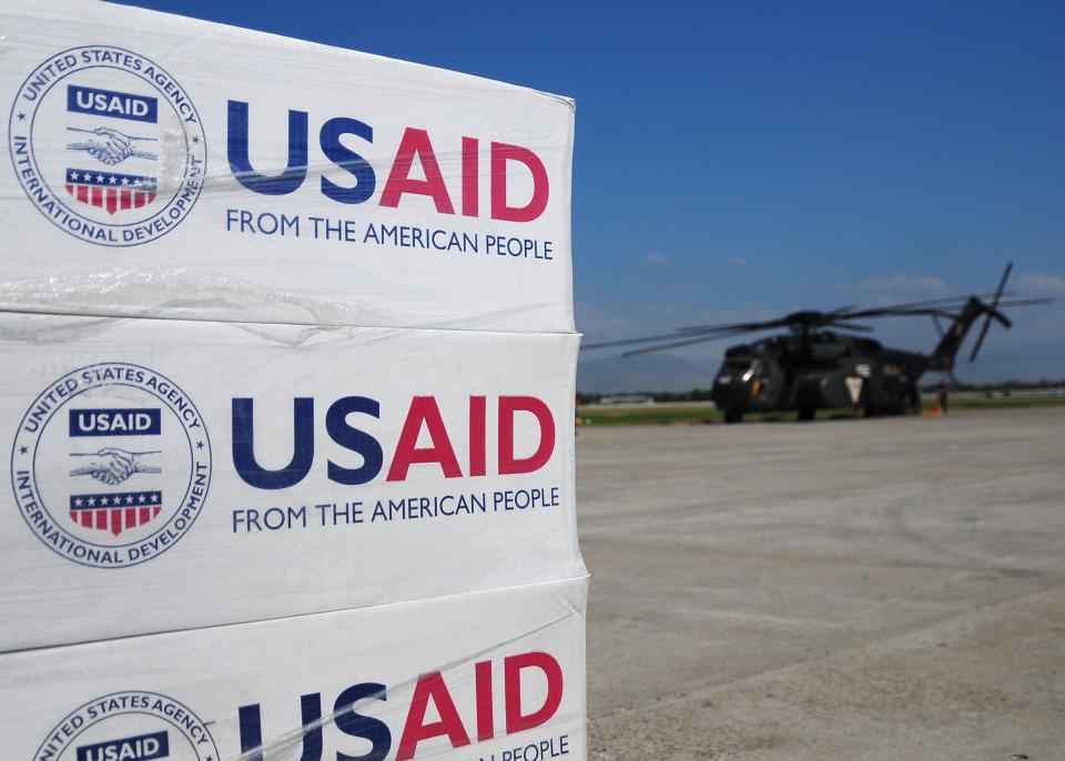 Pallets met USAID logo erop, erachter een helicopter.