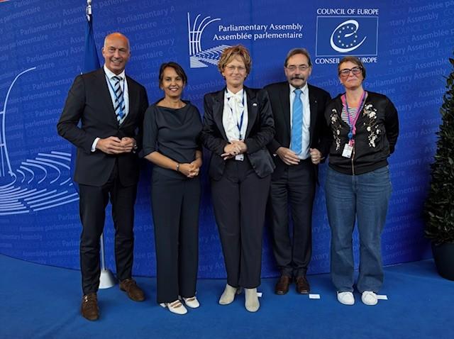 Vijf delegatieleden op een rij poseren voor de foto tegen een donkerblauwe achterrond.