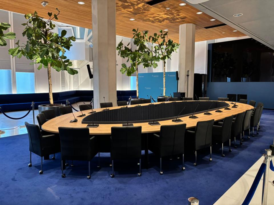 Op een hardblauwe vloerbedekking staat een grote houten ovale tafel met een ruimte in het midden. Er staan zwarte stoelen omheen. Achter de tafel twee bomen in potten.