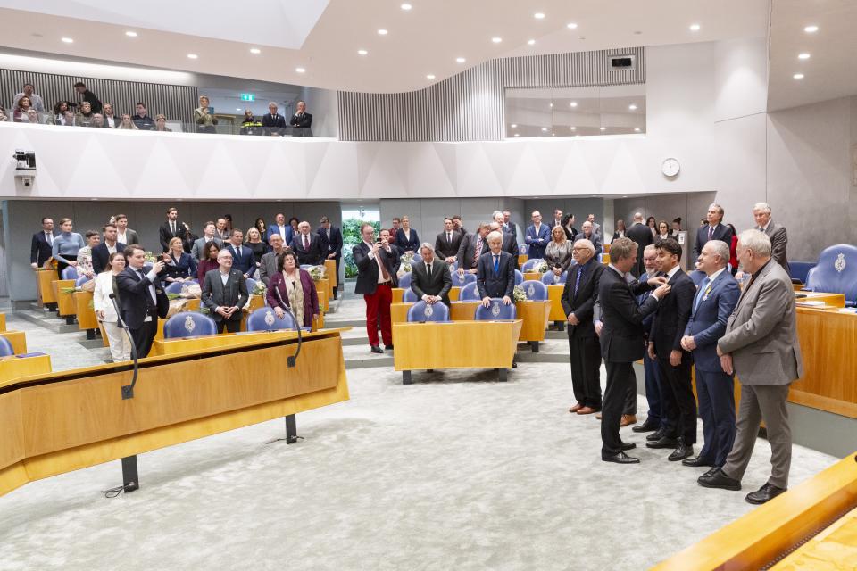 In het midden van de plenaire vergaderzaal staan rechts zes mensen op een rijtje, Kamervoorzitter Martin Bosma speldt hen een voor een een lintje op. 