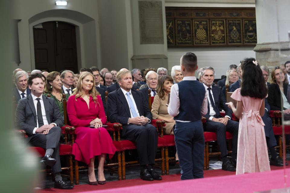 Twee kinderen op de rug gezien, zij bieden een doosje aan aan de leden van het Koninklijk Huis, die op een rij stoelen zitten samen met de Kamervoorzitters en minister-president Dick Schoof. 