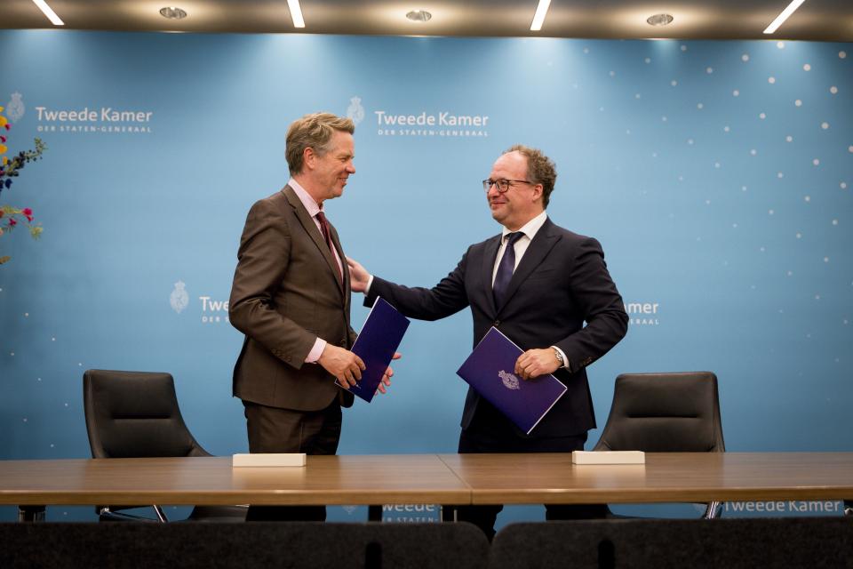 Twee mannen staan achter een tafel tegen een blauwe achterwand. Ze hebben allebei een boekwerk in de hand. 