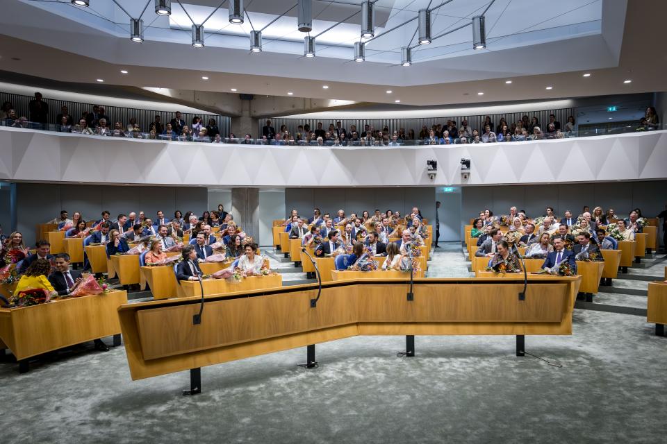 Kamerleden zitten in de plenaire zaal. Er liggen bloemen op de tafels voor ze.