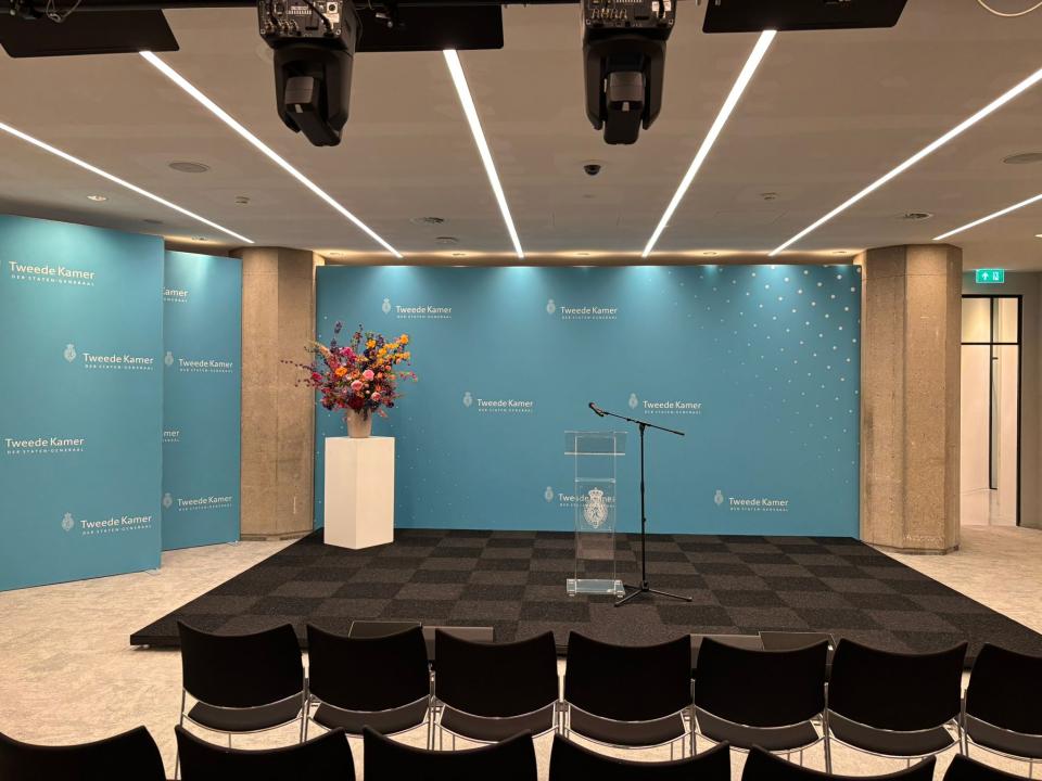 De zaal ingericht voor persconferenties: een blauwe achtergrond, een zwart podium en daarvoor stoelen en aan het plafond camera's. 