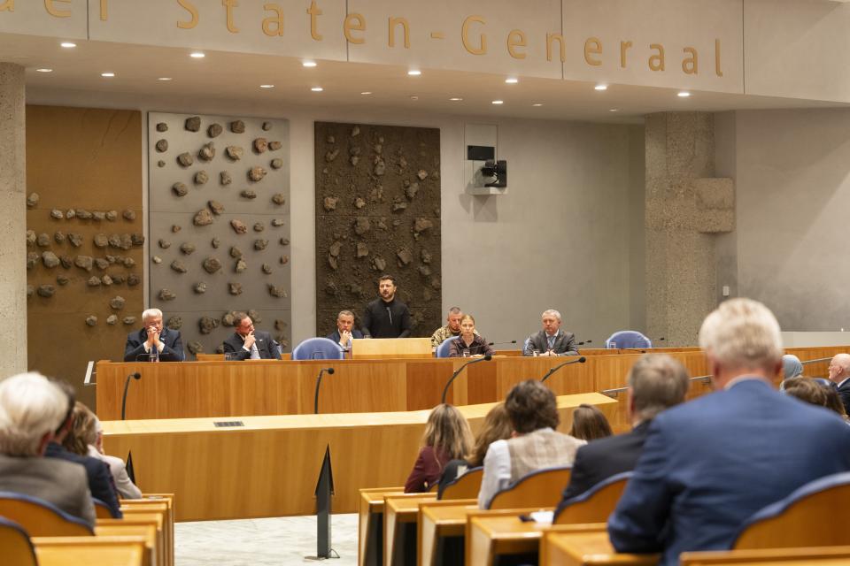 In de plenaire zaal van de Tweede Kamer is in de verte president Zelensky zichtbaar, hij staat achter het spreekgestoelte waar normaal gesproken de minister staat. De Kamerleden zijn op de rug zichtbaar. 