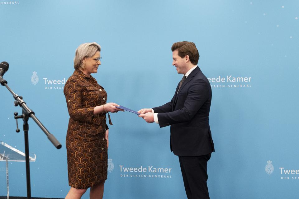 Twee personen staan naar elkaar toe gekeerd voor een lichtblauwe wand met logo's van de Tweede Kamer. Links informateur Letschert, zij geeft een document aan Kamervoorzitter Thom van Campen. 