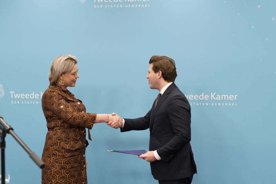 Twee personen staan naar elkaar toe gekeerd voor een lichtblauwe wand met logo's van de Tweede Kamer en schudden elkaar de hand. Links informateur Letschert, rechts Kamervoorzitter Thom van Campen. 