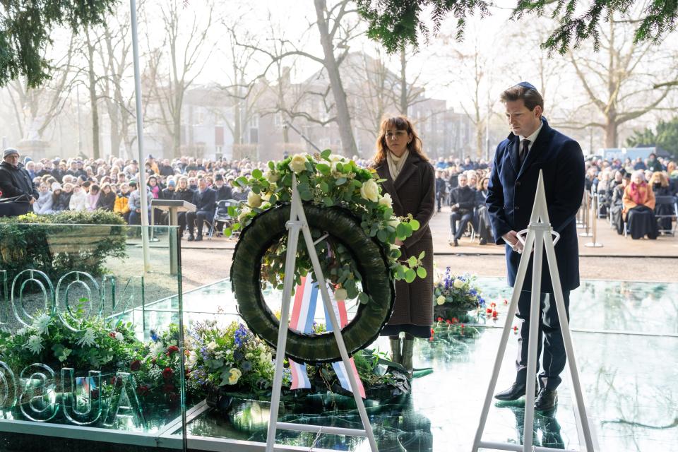 De twee Voorzitters staan voor een krans. Je ziet de krans van de achterkant. Ze staan in de buitenlucht en je ziet een gedeelte van het glazen monument. Op de achtergrond zitten mensen en kijken toe. 