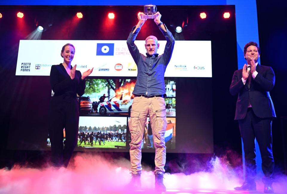 Fotograaf Arie Kievit staat op een podium en houdt de Zilveren Camera trots omhoog. Rechts naast hem applaudisseert Thom van Campen.